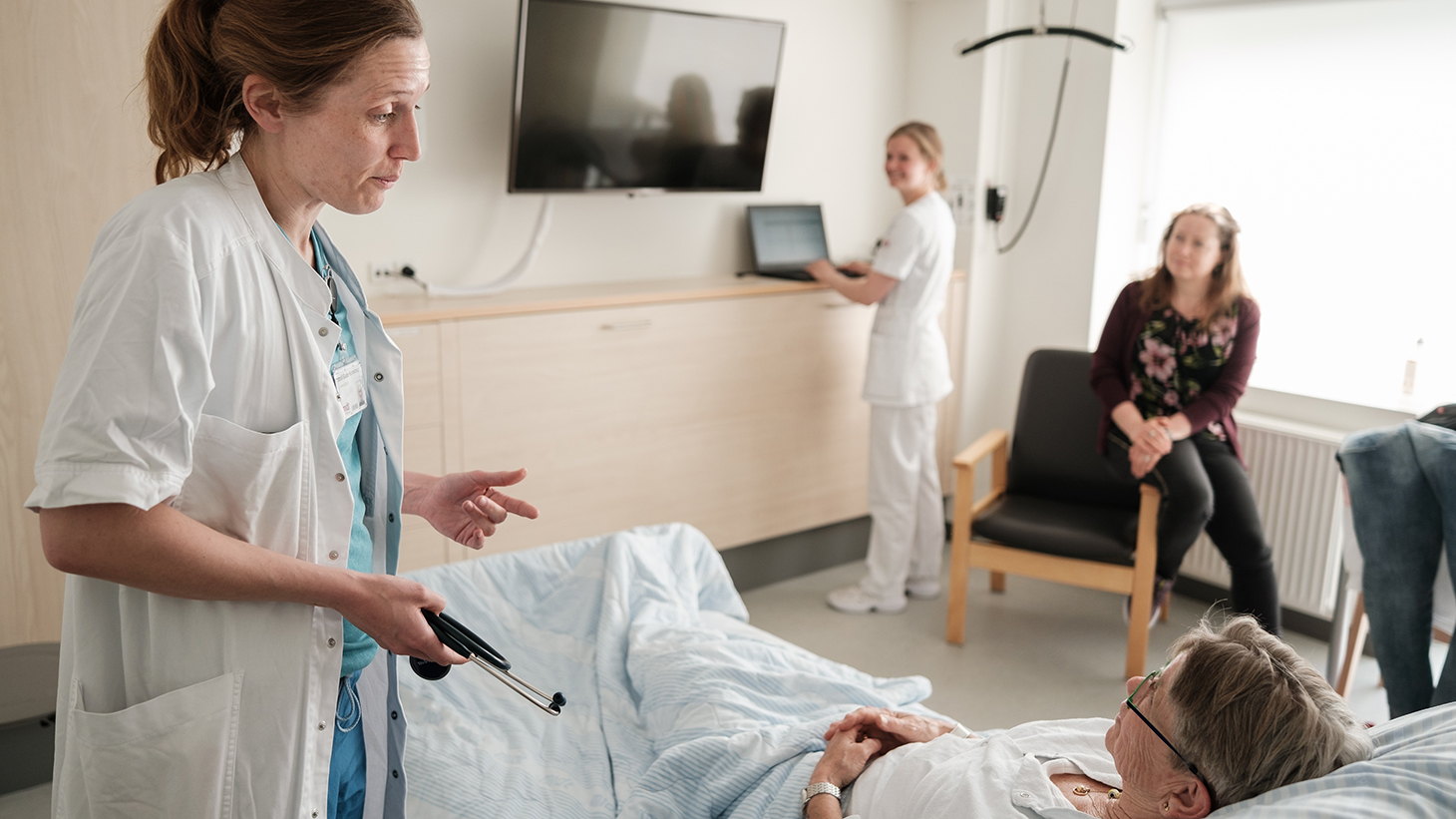 Sundhedsfagligt personale taler med indlagt patient som ligger i hospitalssengen, mens pårørende sidder i en stol ved siden af.